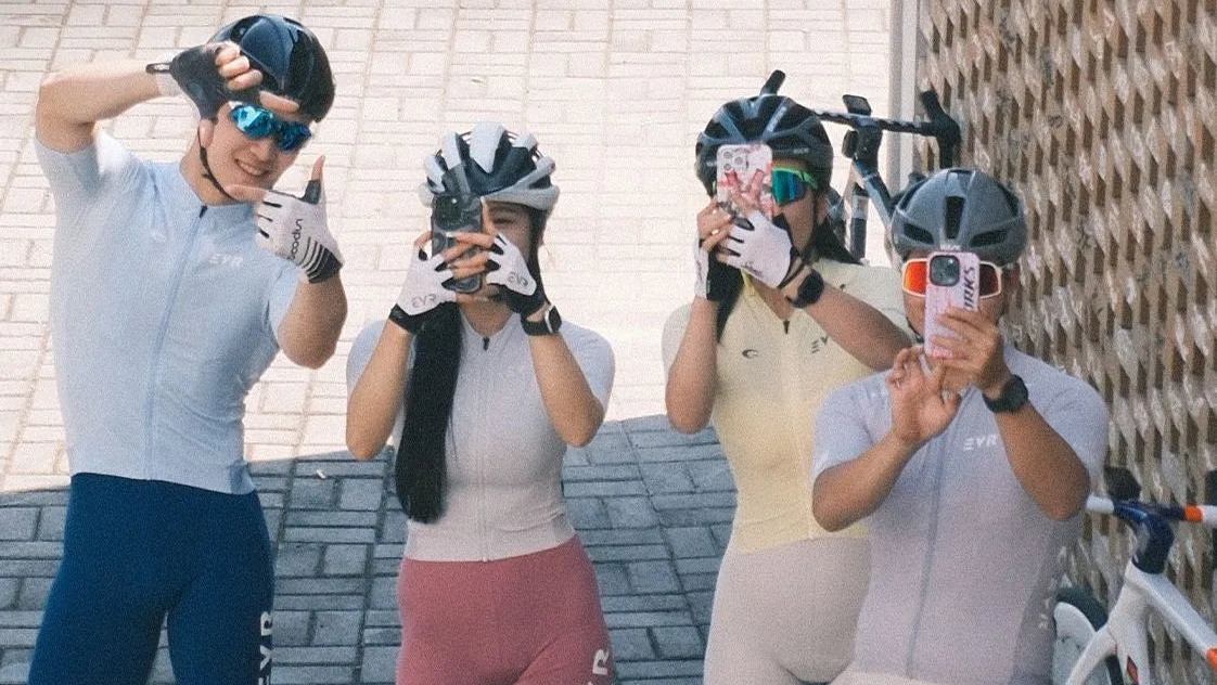 Four cyclists wearing matching cycling jerseys and shorts in different colors (light blue, white, pale yellow, and light gray) pose together on a paved outdoor area next to a bicycle.