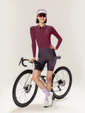 A woman wearing a maroon long-sleeved roam women jersey stands beside a white specialized road bike