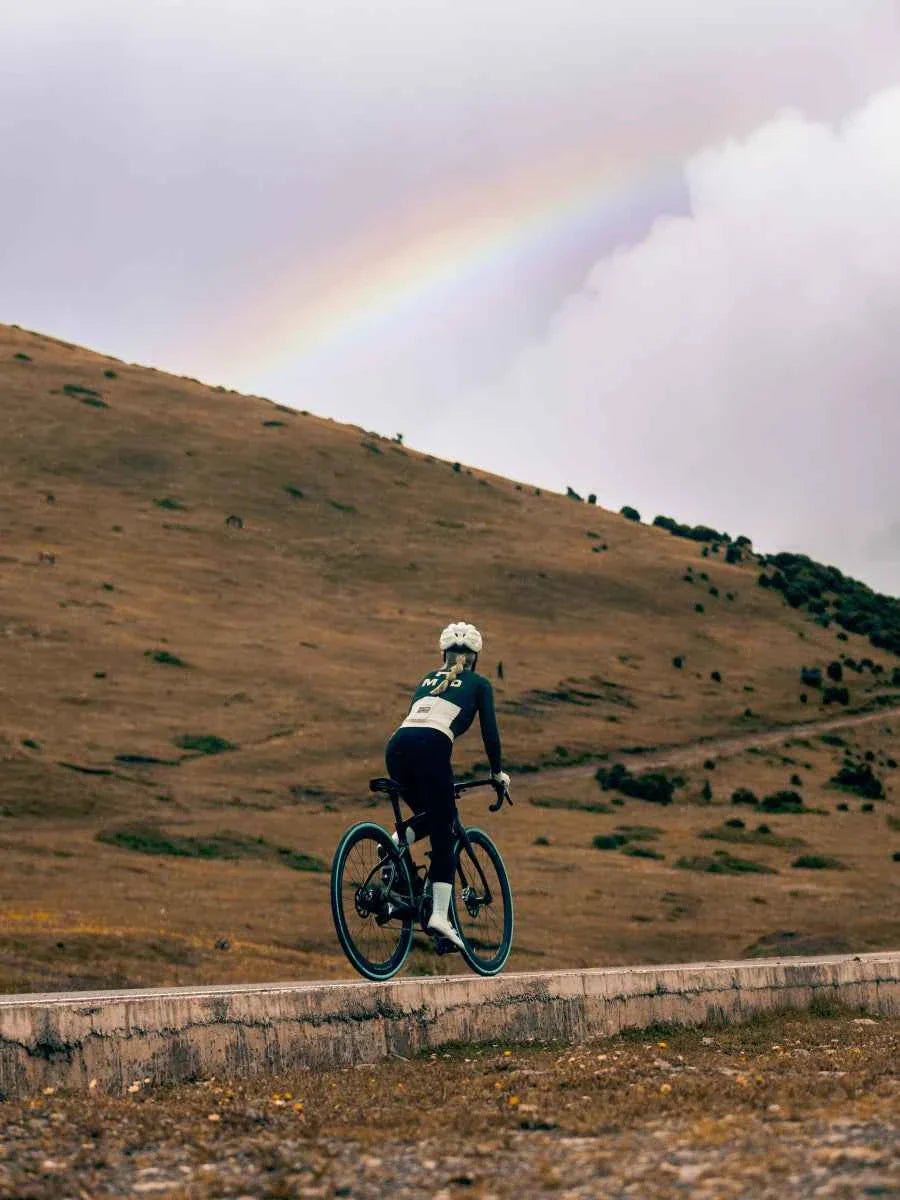 A cyclist wearing a thermal fleeced green freeze women’s jersey rides a road bike under a rainbow