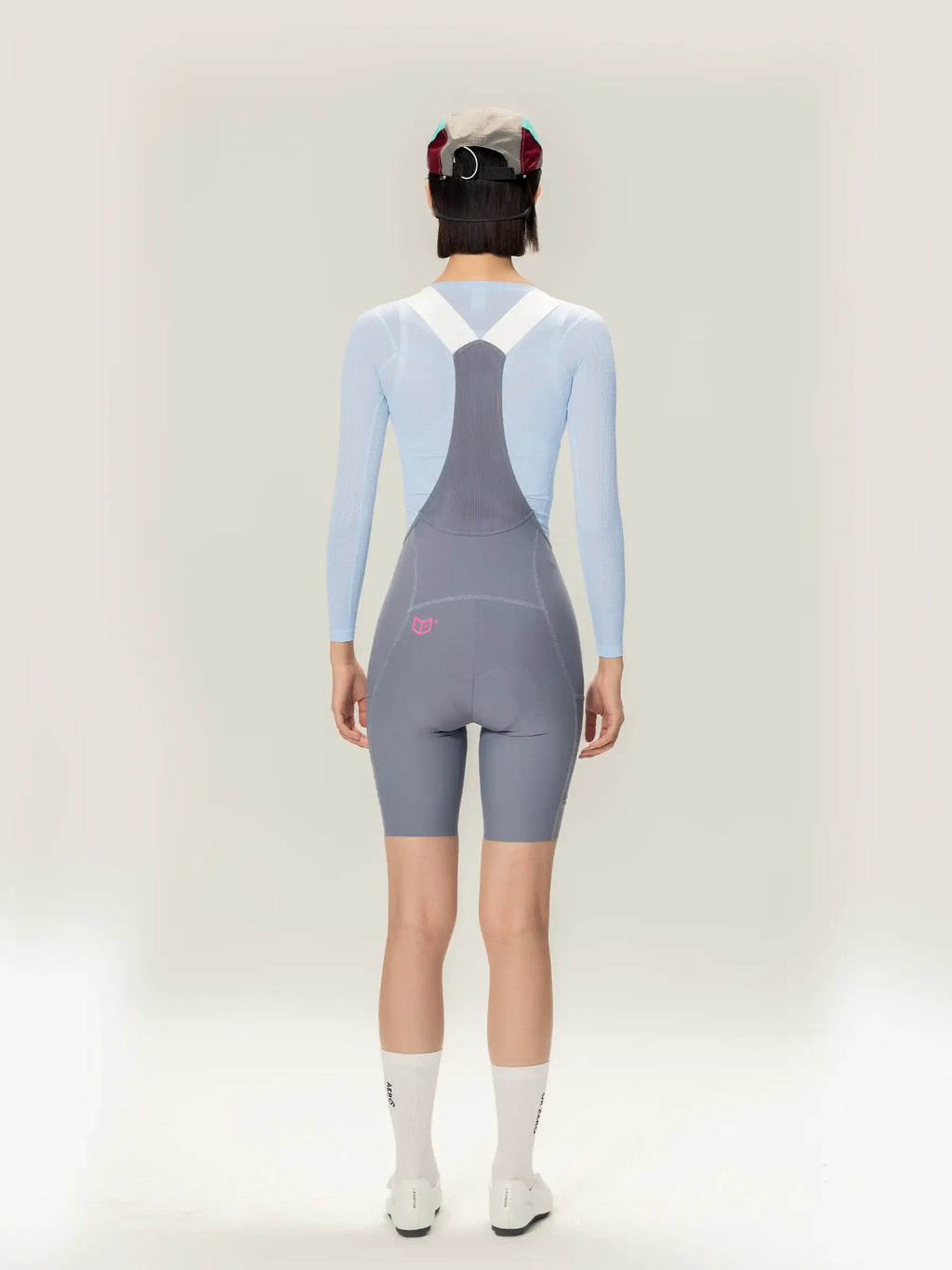 Back view of gray cycling bib shorts with white straps and pink logo paired with lightweave women base layer long sleeve blue featuring airflow mesh for lameda clouds women’s cycling tank use