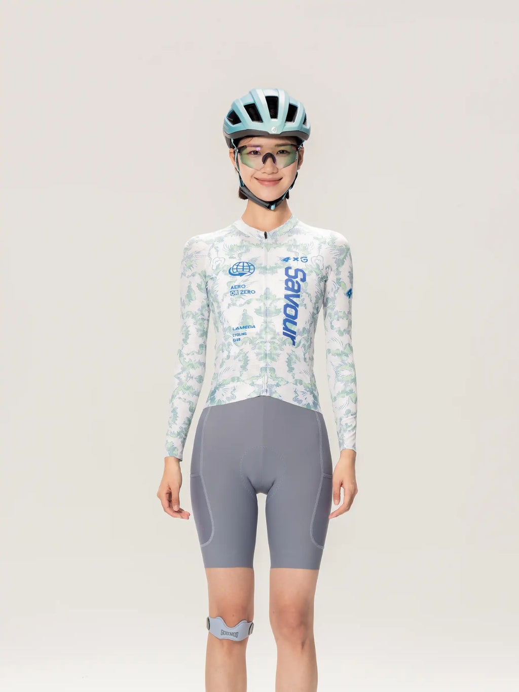 A woman wearing a light blue and white patterned unisex cycling jersey ls from lameda x australian with gray shorts and helmet