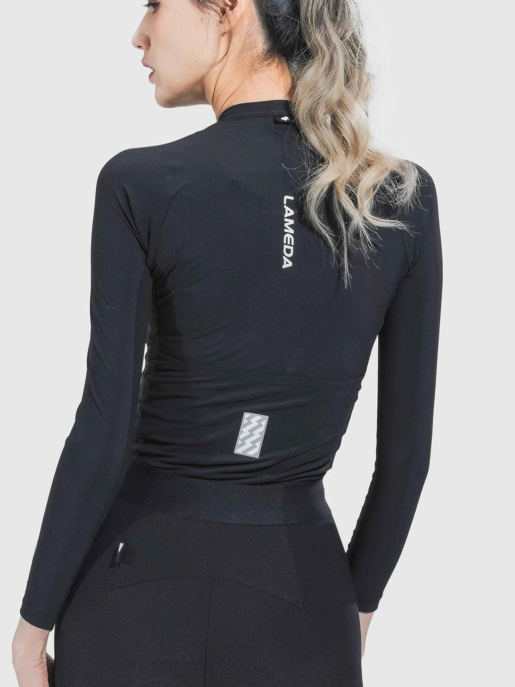 Back view of ripple grace women’s cycling jersey in black with lameda branding and anti-slip hem reflective patch near waist