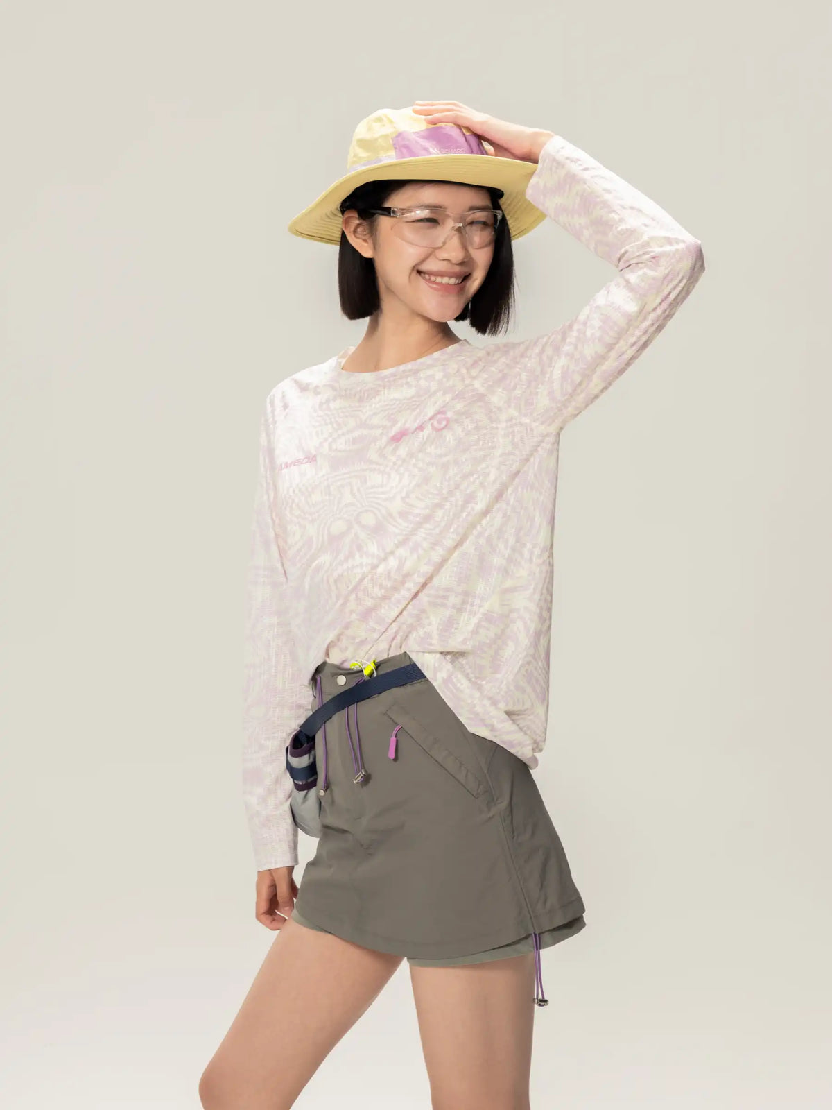 A woman wearing a light yellow hat with purple ribbon and clear glasses modeling a light pink lightweight cycling long-sleeve t-shirt