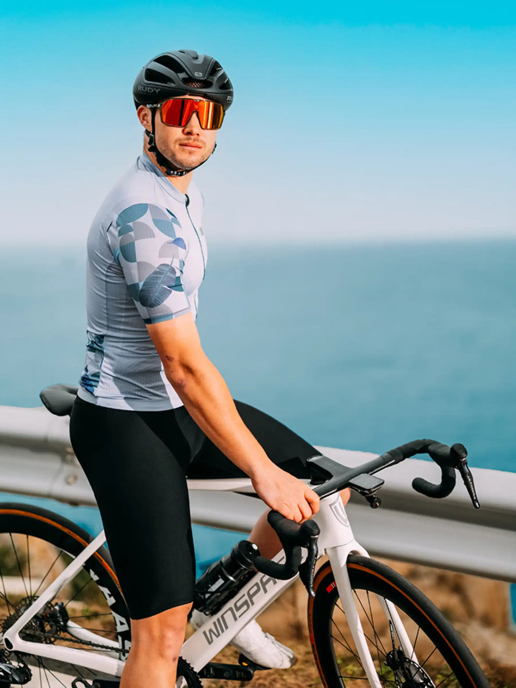 Man wearing a blue relaxed fit cycling jersey riding a white road bike with black handlebars and orange-tinted sunglasses
